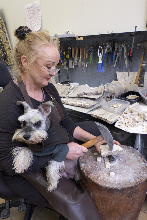 A blonde woman with a small dog on her lap is at work in an artists studio.