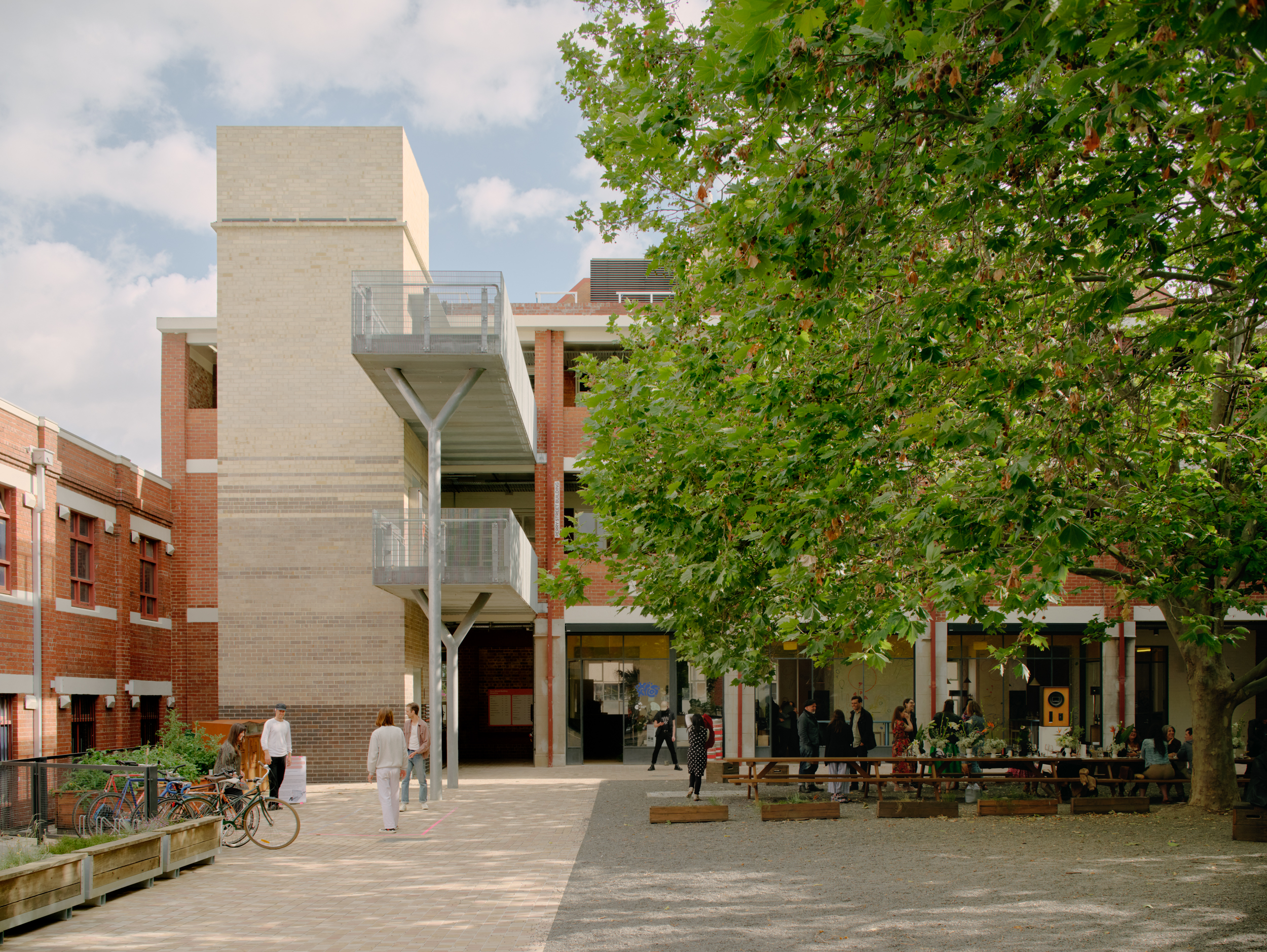 The central courtyard with a leafy tree, and steel walkways and brick facades visible. Some people gather under the tree