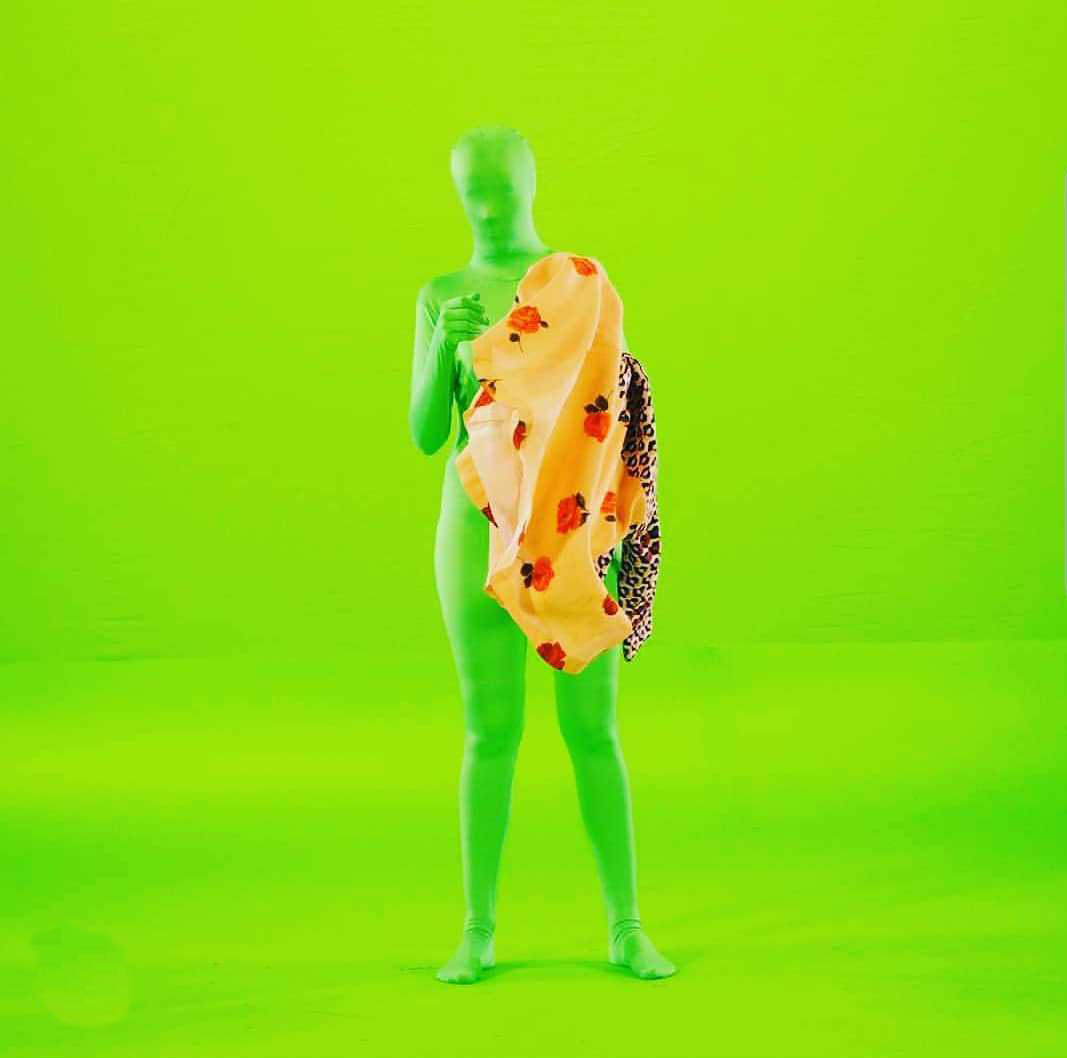 A figure stands in front a bright green screen, wearing a bright green full body suit covering their face. Fabrics are draped over the left side of the body.