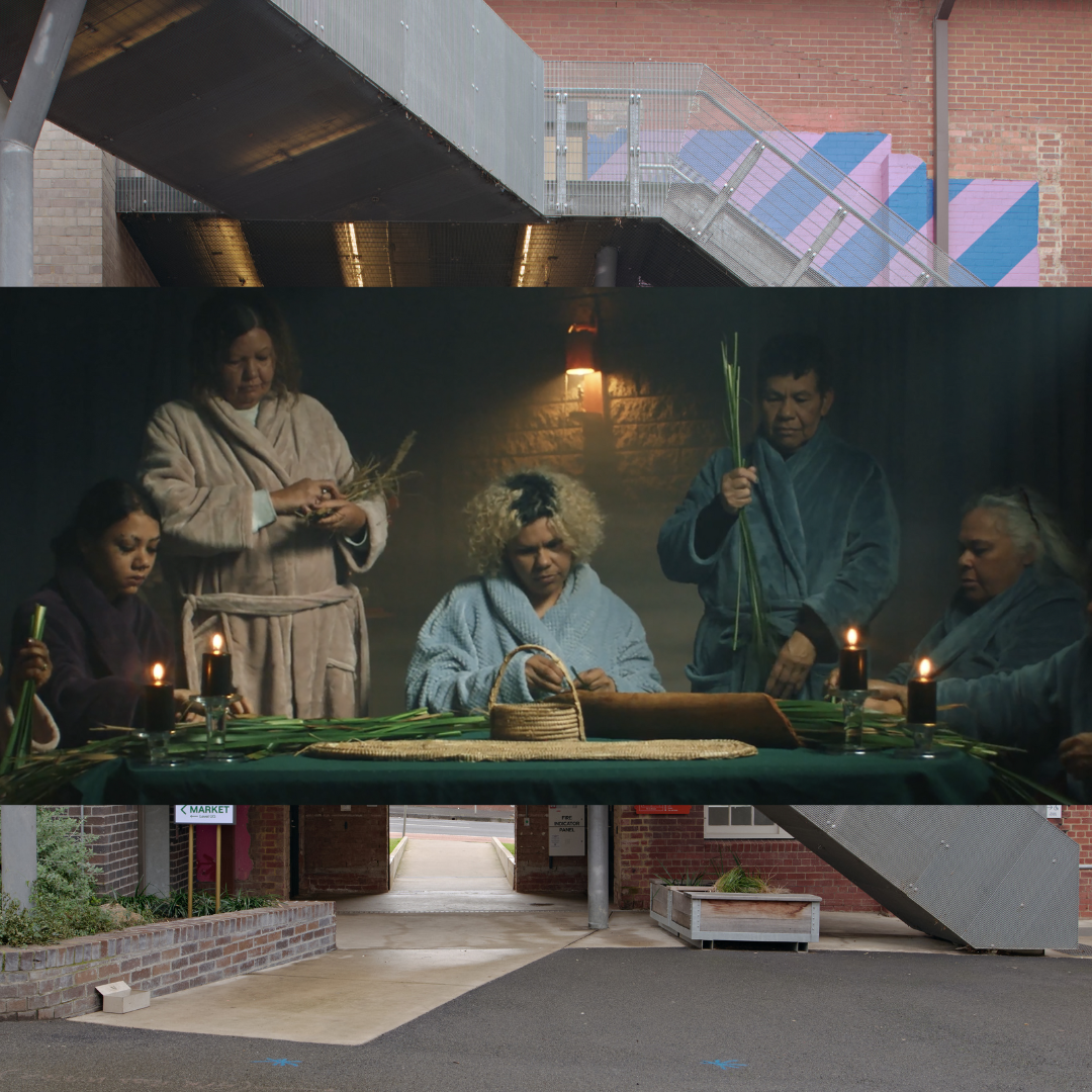 A clip from the video showing a woman in a bathrobe seated before a table and flanked on either side by others in bathrobes carrying leaves and other natural material. The table before them contains baskets, blankets and candles as well as other natural materials. 