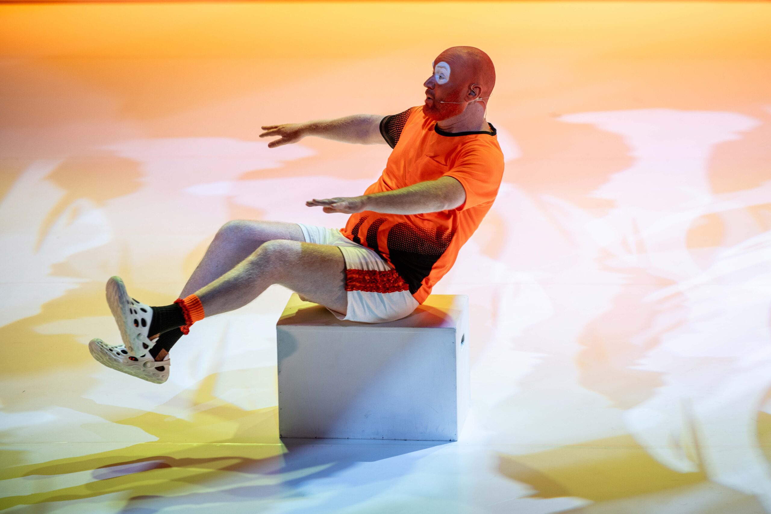 A figure stands in front a bright green screen, wearing a bright green full body suit covering their face. Fabrics are draped over the left side of the body.